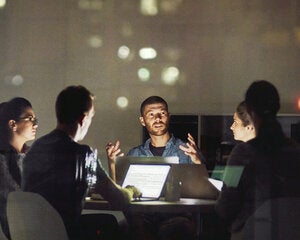 Four professionals collaborating around a lit table with laptops in a dimly lit office at night, with a man gesturing as he speaks to the group.