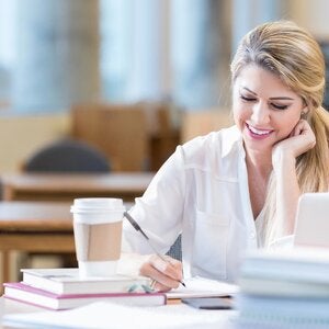 Mid adult female college student concentrates while researching something for an assignment. She is writing in a notebook. An open laptop is on the table.