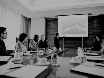 person presenting a graph on a white screen in front of a conference table full of professionals