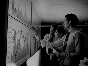 Three business people discuss graphs on screen in meeting room