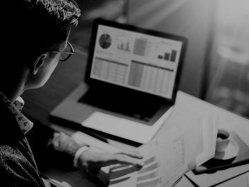 Young finance market analyst in eyeglasses working at sunny office on laptop while sitting at wooden table.Businessman analyze document in his hands.Graphs and diagramm on notebook screen.Blurred.