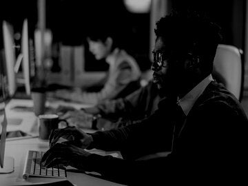 person in front of a computer with another person in the background on a computer