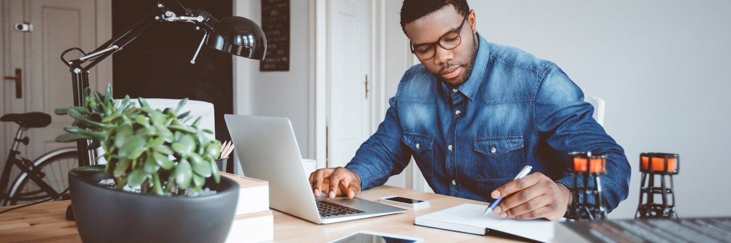 African american man in a home office using laptop and taking notes.